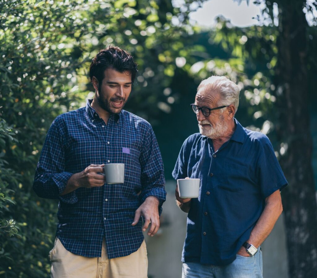 Jeune homme et senior discutant une tasse à la main dans un jardin, image de la cohabitation intergénérationnelle